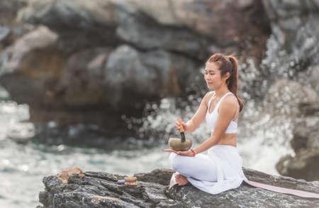 Beautiful Asian woman is practicing yoga meditation by tibetan singing bowl on asana in outdoor with sea wave and mountian rock are background. Healthcare and exercise concept. Yoga Singing Bowl.の写真素材