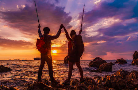 Silhouette of asian couple are hikers with backpacks standing and hands up after success doing...
