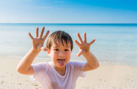 Portrait of happy little boy on summer beach with show sand on hands and face. Child smiling and enjoys on beach with stick out tongue.の写真素材