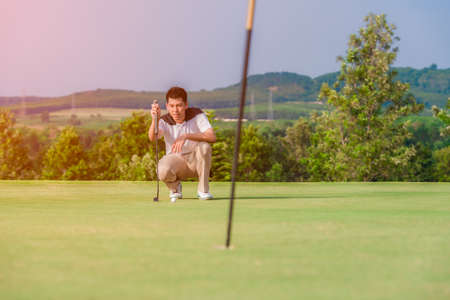 Asian male golfer kneeling holding golf club and checking path line for golf ball on green in outdoor.の写真素材