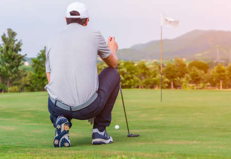 Male golfer kneeling holding golf club and checking path line for golf ball on green.の写真素材