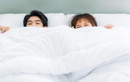 Asian young couple hiding under white blanket on bed with happily in bedroom.の写真素材