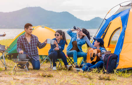 Group of Asian young sitting drinking coffee and playing guitar to happily near lake and the tent camp on outdoors camping. Lifestyle and camping concept.の写真素材