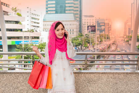 Portrait beautiful happy smiling muslim woman holding shopping bags and talking on smart phone in overpass on city background. Lifestyle concept.の写真素材