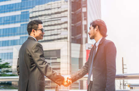 Two happy young businessmen shaking hands in elegant suit on overpass at outdoor on city background. Business partnership concept.の写真素材