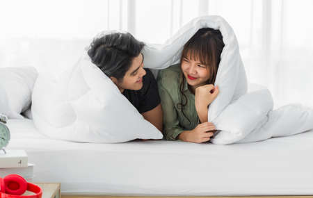 Young couple looking and smiling having fun wrapped in their duvet on white bed in bedroom.の写真素材