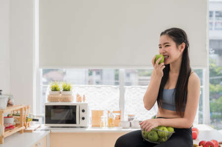 Happy Asian healthy woman in sportswear holding basket and eating green apple in kitchen at apartment. Beautiful sports girl sit smile on cook table and enjoy eat fruit for health after exercise.の写真素材