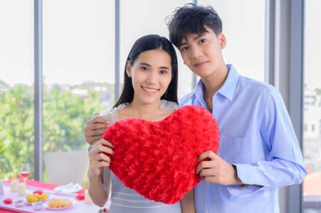 Asian young couple standing smiling and holding red heart pillow in restaurant room on building for romance and celebrating valentine day. Concept love story of young couple in valentine's day.の写真素材