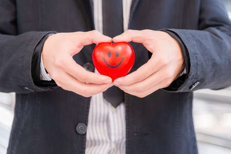 Business man in suit standing holding red heart and making heart shape by hands indoor on city background. Business customer care by heart concept. Valentine day with sweet and romantic moment.の写真素材