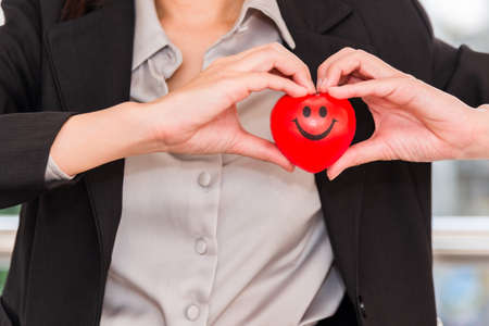 Business woman in suit standing holding red heart and making heart shape by hands indoor on city...