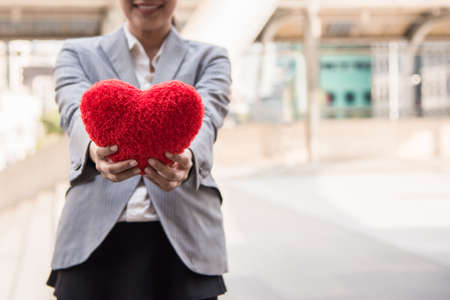 Business woman in suit smiling and standing holding red heart shaped pillow by hands on city background. Business customer care by heart concept. Valentine's day with sweet and romantic moment.の写真素材