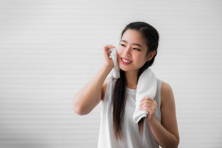 Portrait of happy beautiful asian woman using white towel wiping sweat from her face after exercise in home. Close up of healthy female or sports girl smile and wiping sweat with towel after workout.の写真素材