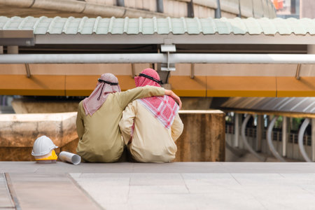 Back view of two Arab engineers sitting on floor and working discussion with safety helmet and blueprint in construction site. Saudi man worker and friend sit hugging the neck talking while break time.の写真素材