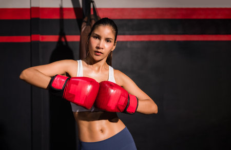 Portrait of confident Asian woman in sportswear wear red boxing gloves with workout and pose boxing show punch in gym. Young female boxer exercise and training by boxing for healthy in fitness center.の写真素材