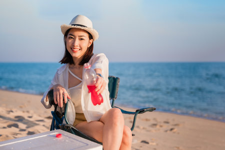 Happy beautiful Asian woman sitting on picnic chair wear sunglasses and hat holding red beverage bottle on beach in outdoor vacation summer. Young female smile relax and drink cocktail travel camping.の写真素材