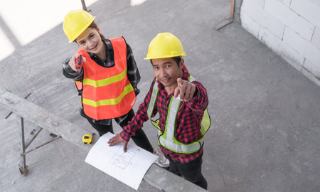 Top view of two Asian civil engineer wear safety hardhat stand look blueprint and point for check infrastructure in construction site. Female architect and male contractor working together on projects.の写真素材