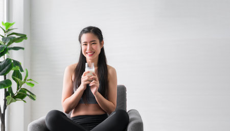 Happy Asian sports girl in sportswear sitting on chair and holding pure milk from glass after exercise in home. Healthy woman smile and drinking milkshake protein. Healthcare and weight loss concept.の写真素材