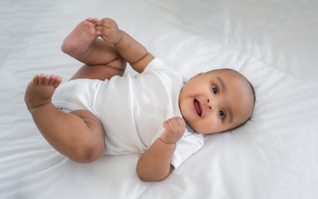 Portrait of happy African American mixed race cute girl baby lying down on white bed and look on camera in a bedroom at home. Top view of little newborn infant smiling laughing and lying on duvet.の写真素材