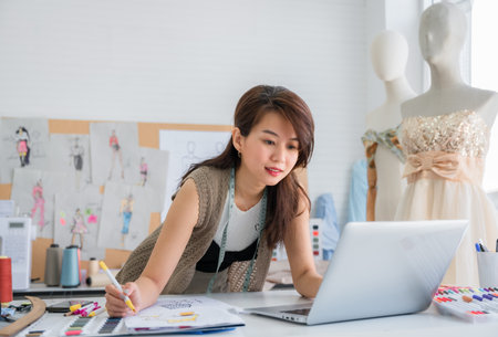 Happy Asian woman dressmaker or designer seamstress tailor with a tape measure using laptop and sketches design for trendy fashion in the studio office. Attractive female stylist with mannequin dummy.の写真素材