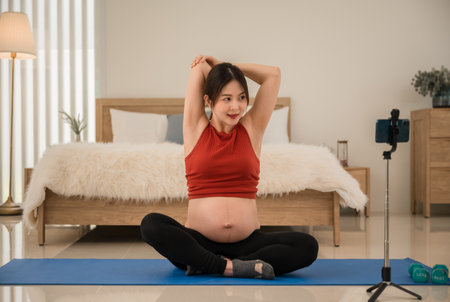 Happy Asian pregnant woman in sportswear sitting on yoga mat stretching warm up and exercise from looking online class by smartphone in the room at home. Healthcare of mother and unborn baby concept.の写真素材