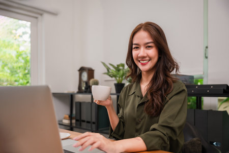 Portrait of Happy beautiful Asian freelancer businesswoman sitting smiling and using a computer laptop for working and holding a coffee cup for drink with looking at camera in a private room office.の写真素材