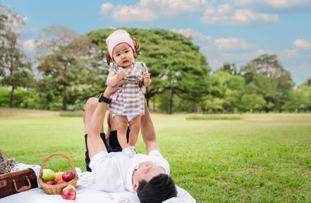 Portrait of happy cute little Asian baby girl and her father holding and playing together having fun and enjoying while picnic on lawn outdoors in park. Cheerful adorable small daughter with her dad.の写真素材