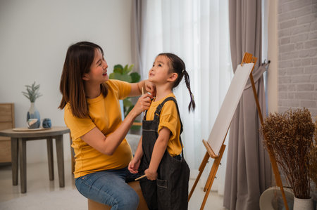 Asian pregnant mother dressing and putting on an apron on her little artist daughter before painting with happiness together in the living room at home. Pregnant mom and her child girl bonding concept.の写真素材