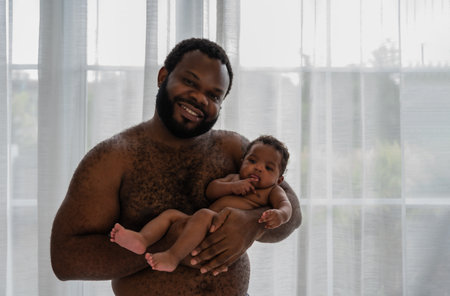 Portrait of happy African father shirtless standing smiling and holding his newborn baby son in his arms with love and affection with looking at camera in the bedroom at home. Father's Day concept.の写真素材