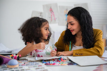 Cute little African girl with an afro hairstyle sitting smiling and learning sewing from her beautiful mother dressmaker and designer together happiness in the fashion studio workplace.の写真素材