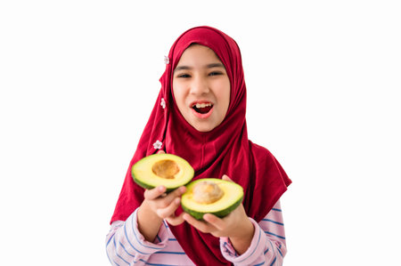 Portrait of happy cute Muslim child girl in hijab standing smiling and holding a fresh avocado fruit...