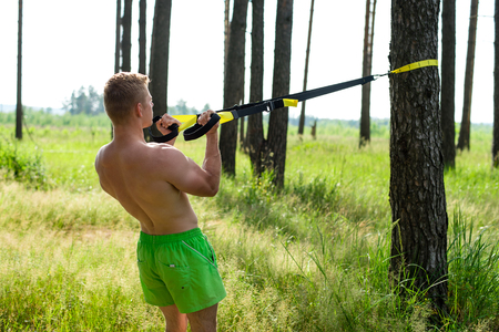 Male athlete excellent training trx, fresh air in nature in the summer in the forest, feel your strength, motivation, close-up. Exercise of the biceps muscles. A sunny day. Stamina of a man. Against the background of trees.の写真素材
