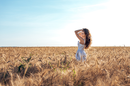 Beautiful happy woman in a field, sunny afternoon, white dress. Brunette hair, tanned skin, concept of enjoying nature. Happy smiling. Harmony with wheat.の写真素材