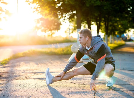 A male coach kneads his legs before training with headphones. Runner in morning in the park. Listens to music, confident look. Summer lifestyle, motivation is strong. Outdoors in the city.の写真素材