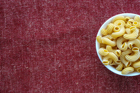 cavatappi pasta in a white bowl on a red brown cloth burlap background on the side. With space for...