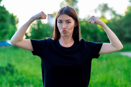 Brunette girl in summer outdoor. Black T-shirts on a green meadow. He shows his biceps with gestures. Emotion is a joke power.の写真素材
