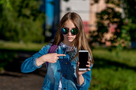 Little girl schoolgirl in sunglasses. Summer in nature. In his hands holds a smartphone. A finger points to the phone. The concept is a new gadget app. Emotion focus concentration.の写真素材