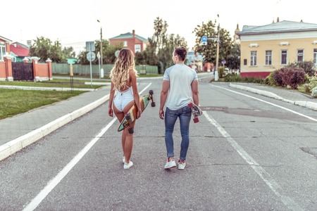 Young couple view from the back, go through the city in the summer. Skateboards, longboards, walk in fresh air are kept in ruqas. A girl in a white body bathing suit, guy in a t-shirt and jeans.の写真素材