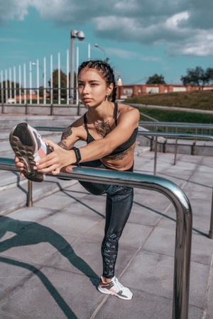 The girl with tattoos in sportswear does a warm-up before gymnastics. For morning run. Against the background of the stairs, in summer in city. Sports and healthy lifestyle.の写真素材