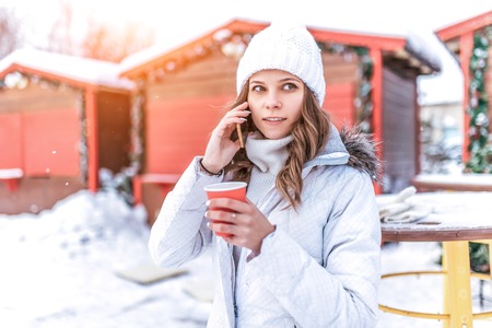 Beautiful and young girl in a white hat in winter sunny day. In his hands holding a cup of hot tea...