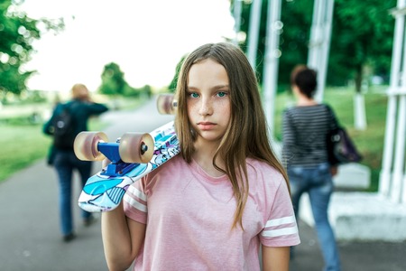 Teenager girl in the summer in the city, sad stands in the park, upset, holding a skateboard on her shoulder. Long beautiful hair. Concept of difficult teenagers. Freckles on the face.の写真素材