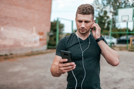 Young guy athlete man, serious look screen of smartphone, talking on video call. Summer in city park on background of sports field. In casual clothes with headphones, listening music.の写真素材