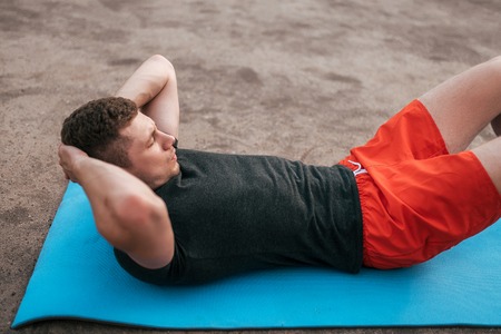 Young guy on rug. Trains twisting, abdominal muscles. Strengthening abdominal muscles. Raise torso. The concept of a healthy lifestyle or hobby. Man in the summer on the sports ground in city.の写真素材