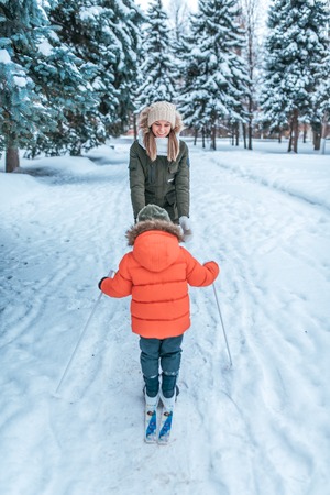 4-6 years old boy rides and walks on toy skiing with young mother. Winter forest drifts and wood drift on background. First steps in sport active lifestyle of child. Care and support parents winter.の写真素材