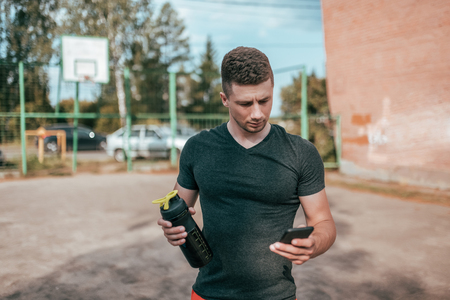 Male athlete, summer sports field in hand smartphone, writes message to Internet, an app pulse and workout activity. Active lifestyle fitness workout. In his hand bottle of shaker with water.の写真素材