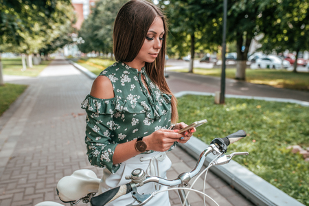 Beautiful girl with the phone watching videos on the Internet, social networking, online, biking in the summer in the city. A woman listens to music on headphones.の写真素材