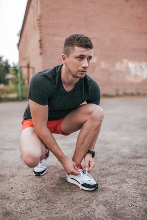 Athlete man crouched, tying shoelaces on sneakers. In summer on the sports ground in the city. Trainer guy in the watch tracker bracelet after jogging. Workout outdoor lifestyle.の写真素材