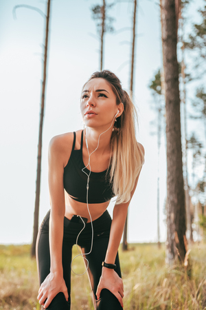Beautiful girl woman athlete summer forest. Listening music headphones phone. Tired, resting after running shortness deep breath. Motivation for running woods. Emotion endurance strength confidence.の写真素材