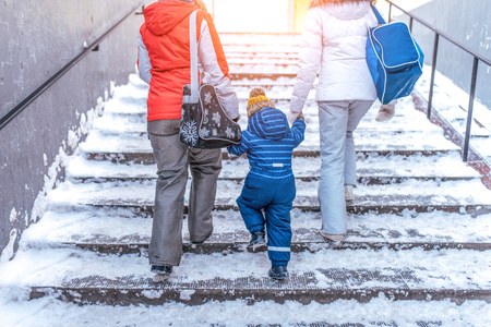 Parents mom and girlfriend girl hold hand little boy son 4-5 years old, go up stairs in transition, winter city backdrop snow mud. View from back, in hands of bag with gangs, will return after rink.の写真素材