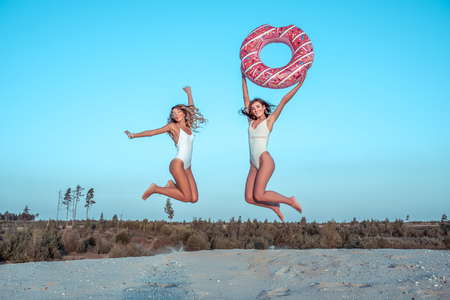 Two girls girlfriends jumping having fun playing with inflatable circle, white sand, sea quarry, lake beach. Fashion lifestyle. Tanned figure. Active lifestyle woman. Holiday birthday fun vacationの写真素材