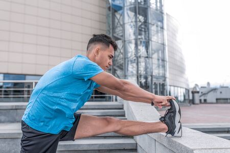 Athletic man running, warming up leg muscles, before training, in summer in the city, sportswear t-shirt shorts, sneakers, fitness in the morning on a runの写真素材